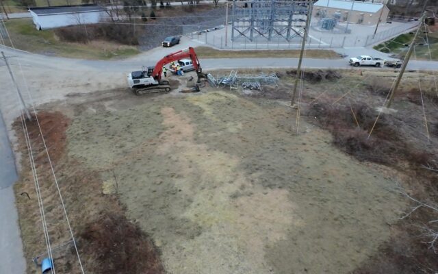 Red excavator, vehicles, and workers near substation on grass.