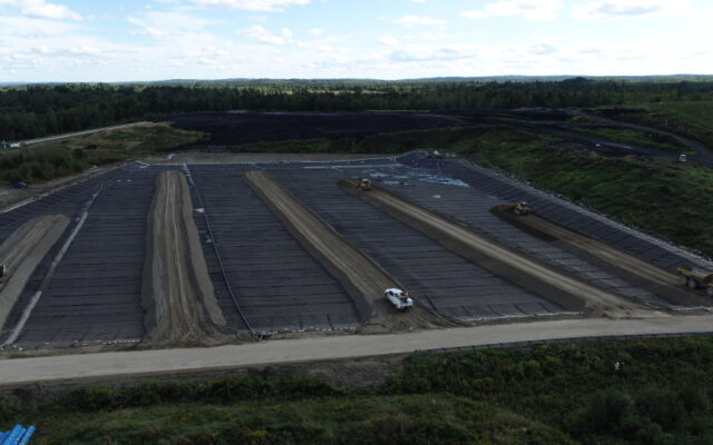 Vehicles work on soil mounds with black liners, trees nearby.