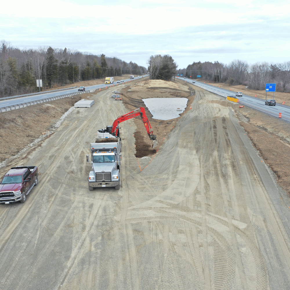 Excavator loads dump truck by highway; pickup and cones nearby.