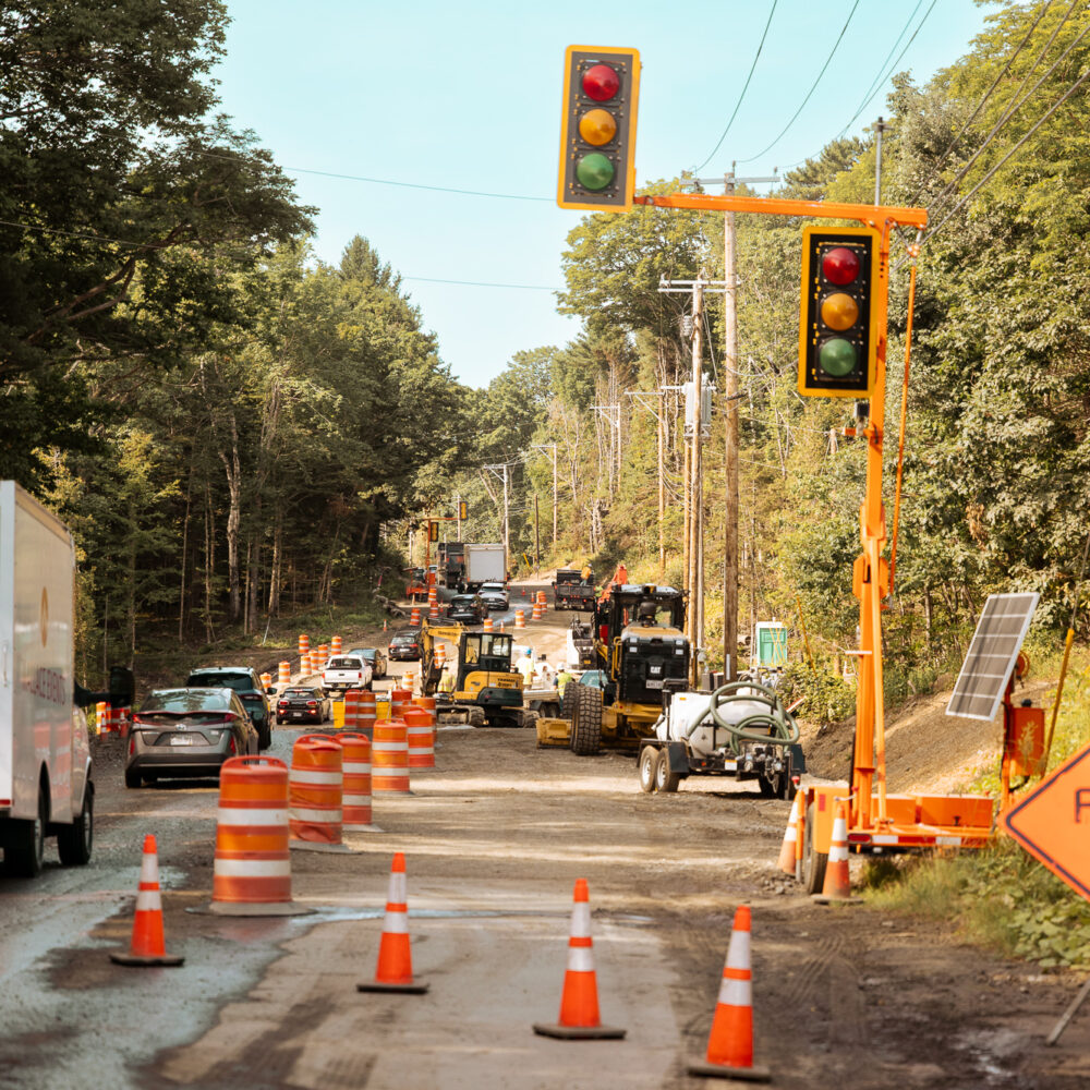 Road construction with traffic, cones, machinery, signs, and trees.