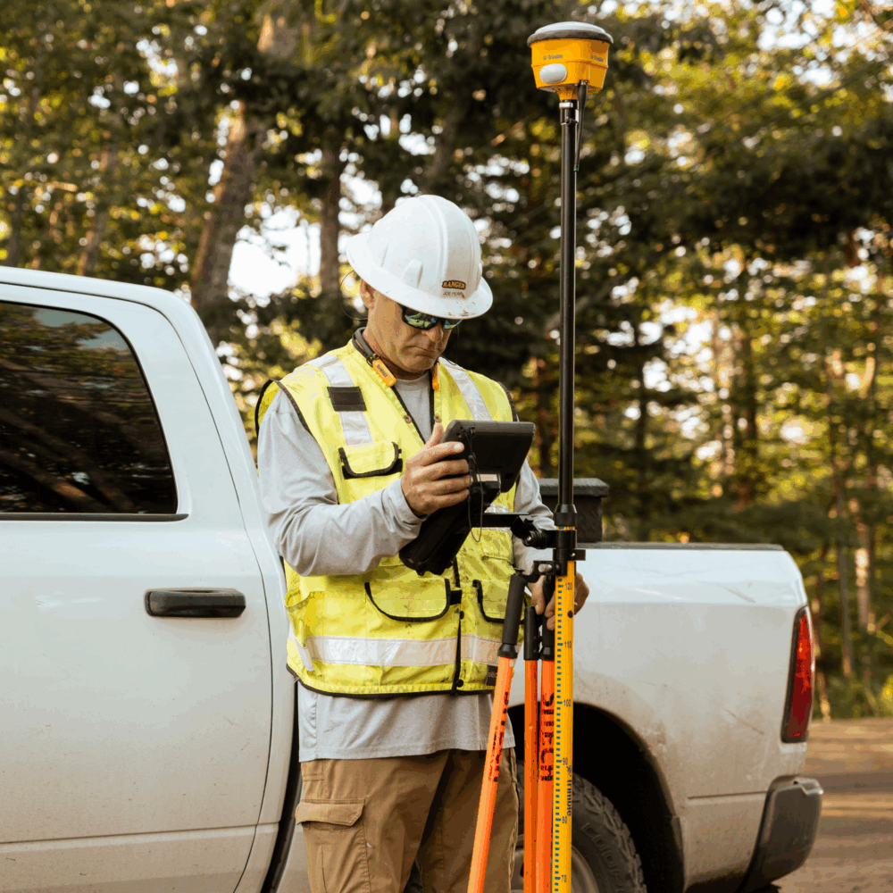 Worker in safety gear surveys near white pickup on dirt road.