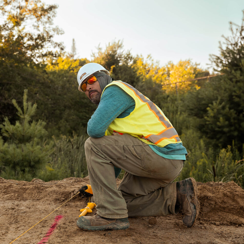 Worker in safety gear kneels on dirt path, measuring tape in hand.