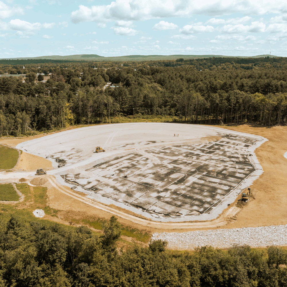 The image depicts a large, cleared area of land with construction machinery operating on the surface, surrounded by forested hills and a partly cloudy sky.