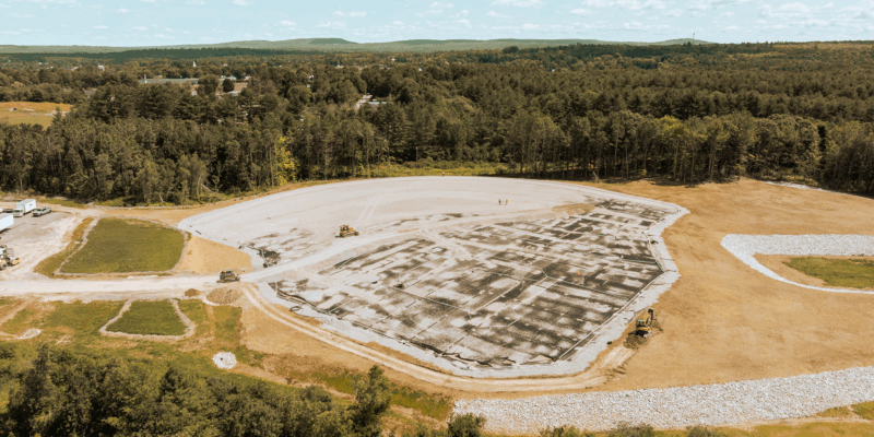 The image depicts a large, cleared area of land with construction machinery operating on the surface, surrounded by forested hills and a partly cloudy sky.
