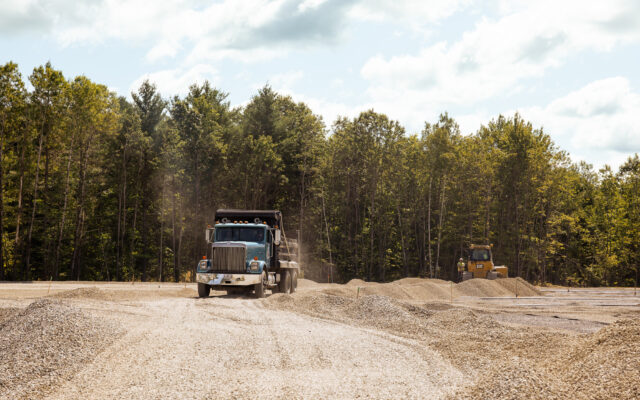 A construction site featuring two heavy trucks, one actively transporting gravel while another operates in the background among piles of aggregate material.