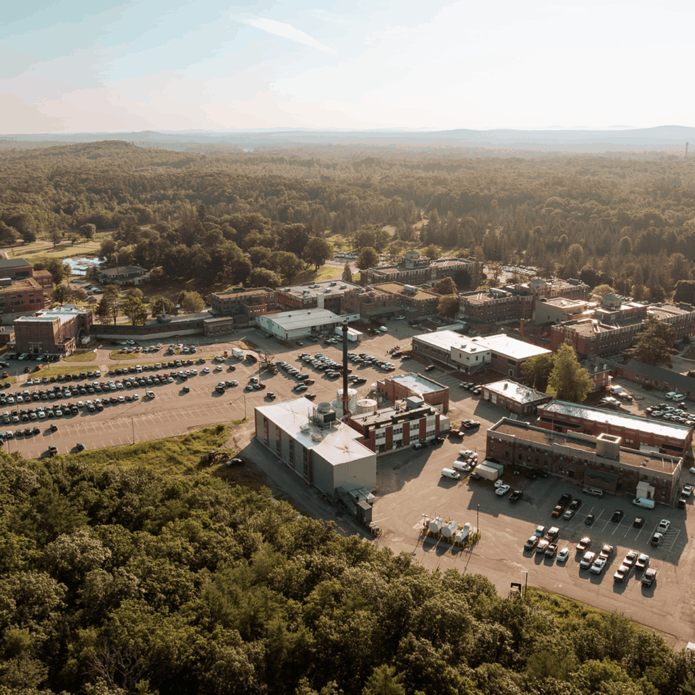 The image shows an aerial view of a complex with multiple buildings and a parking area surrounded by trees and hills in the background.
