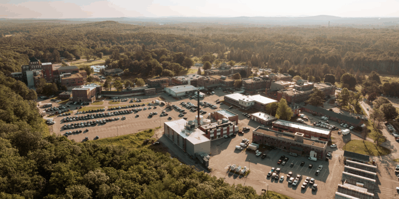 The image shows an aerial view of a complex with multiple buildings and a parking area surrounded by trees and hills in the background.