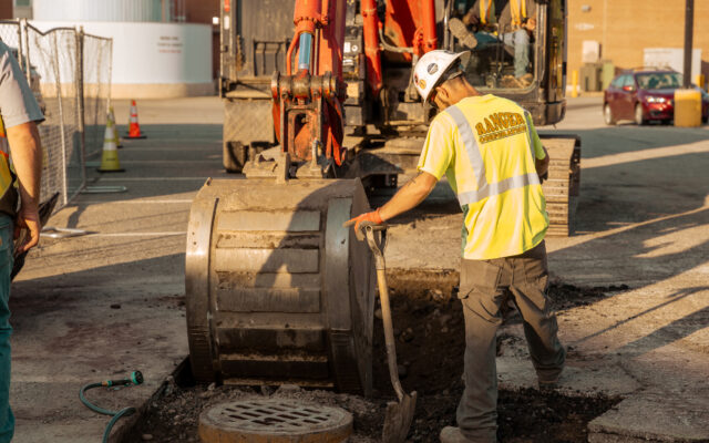 A construction worker in a safety vest is excavating a trench at a site while heavy machinery operates nearby.