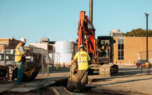 A construction worker in safety gear is reaching into a trench at a worksite, while heavy machinery is positioned nearby.