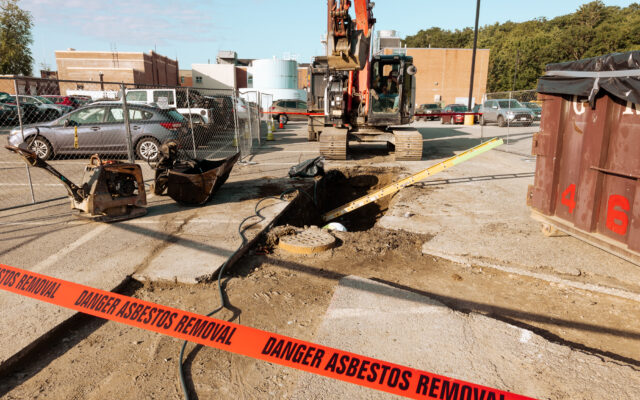 The image shows a construction site with an excavator, a large hole in the ground, and caution tape indicating a danger of asbestos removal.