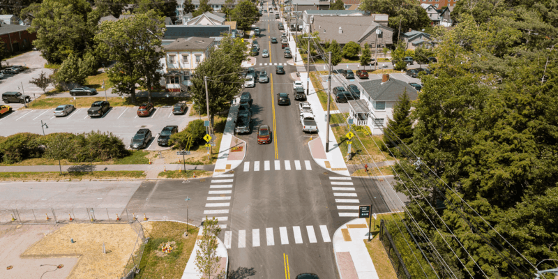 The image captures an aerial view of a busy street lined with trees, houses, and parked cars, showcasing a mix of residential and commercial areas.