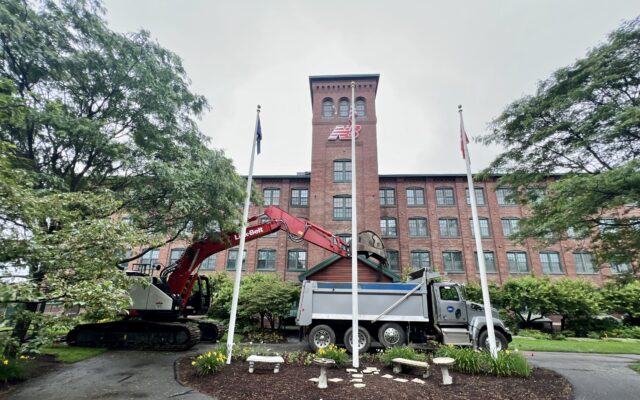 Red excavator loads debris into white dump truck by brick building.