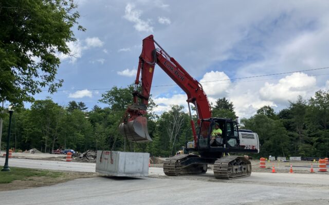 Red excavator lifts concrete barrier at construction site.