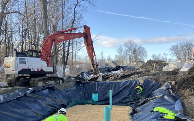Two workers spread gravel in a trench near a parked excavator.