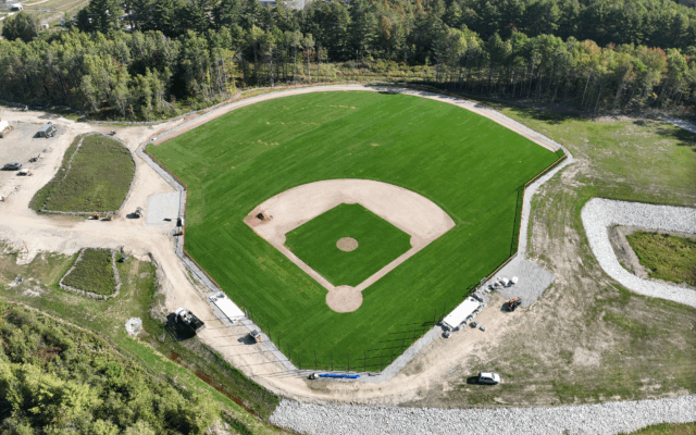 Empty baseball field with trees, gravel roads, and parked vehicles.