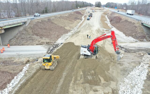 Excavator and bulldozer grade road under overpass; workers nearby.