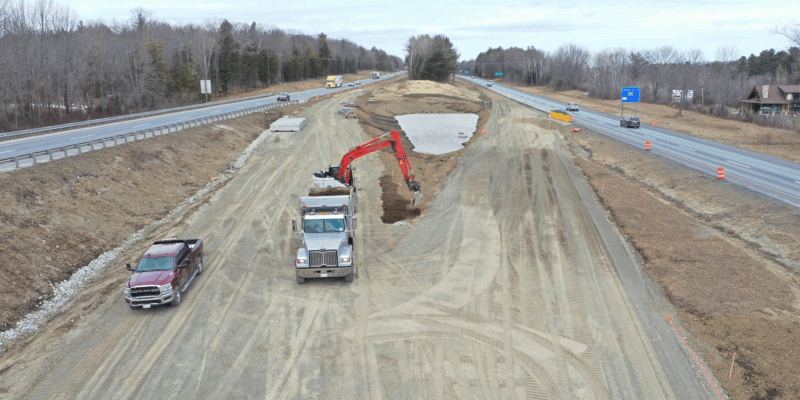 Excavator loads dump truck by highway; pickup and cones nearby.
