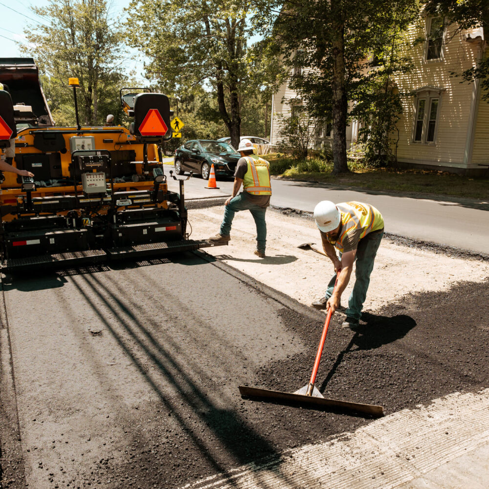 Workers repave a road with asphalt near houses on a sunny day.