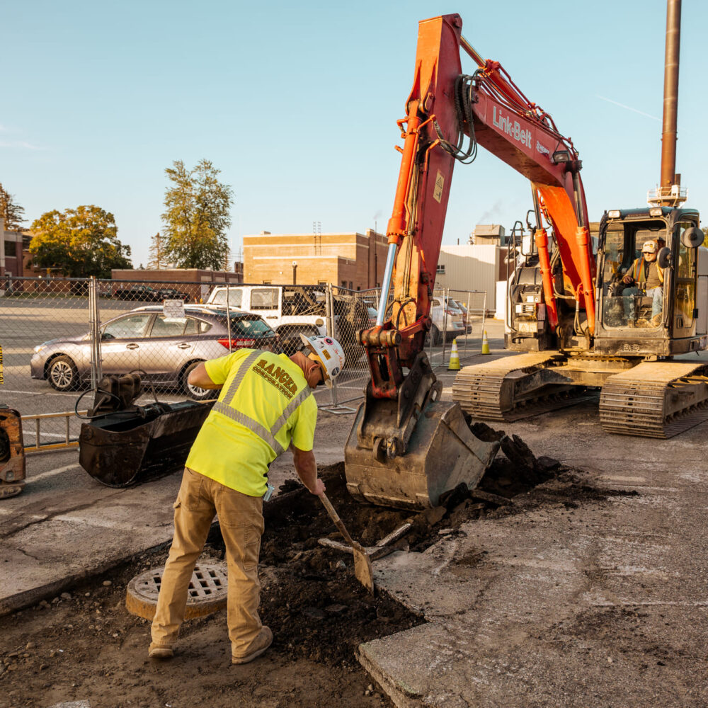 Worker shoveling debris as excavator digs in a parking lot.
