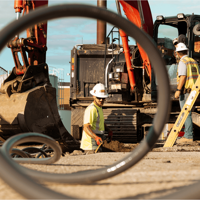 Workers in safety gear operate machinery, seen through circular frame.