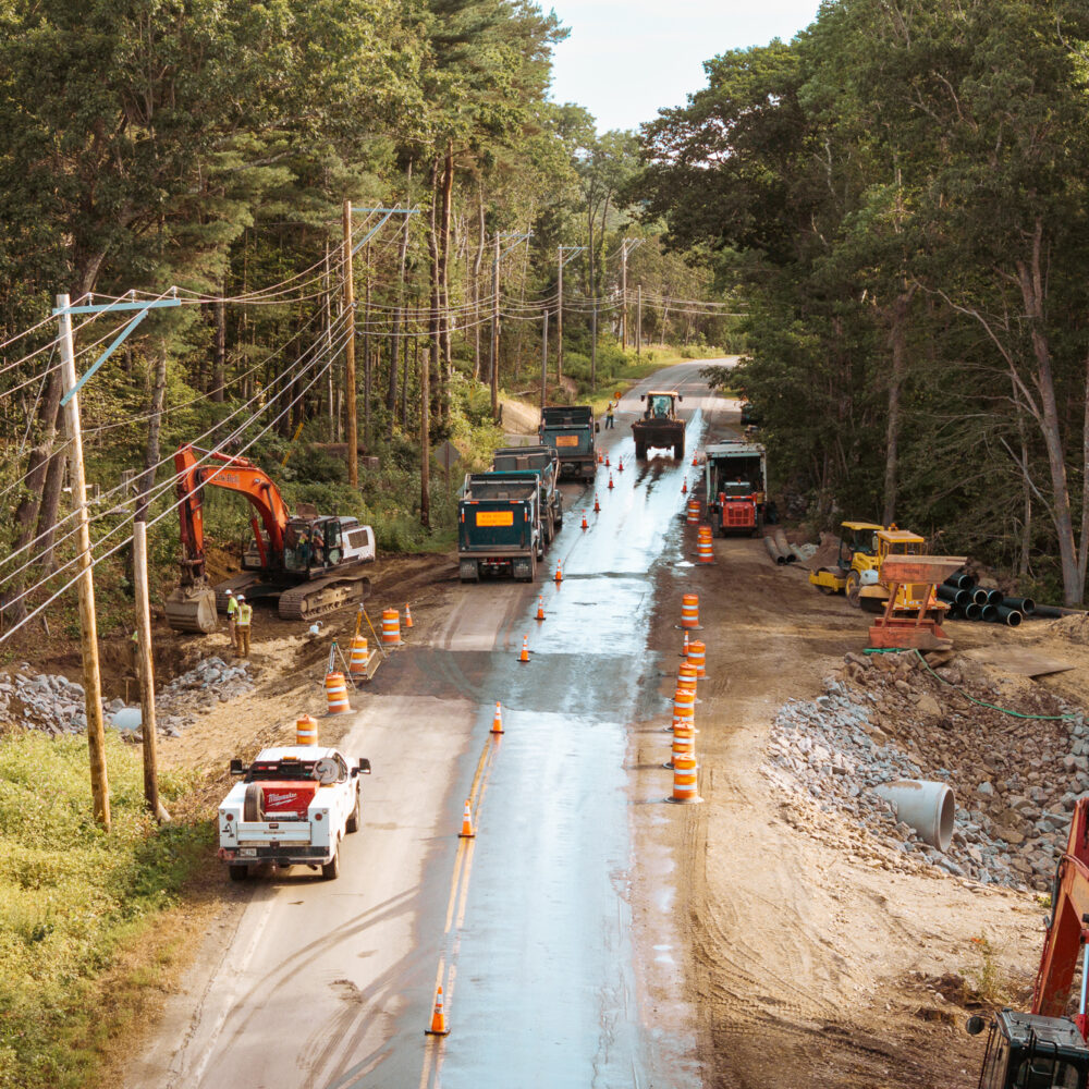 Rural roadwork with vehicles, cones, workers, trees, and poles.