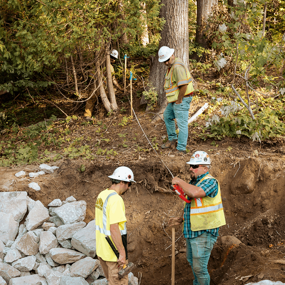 Three construction workers are collaborating on a project in a wooded area, examining a dig site with rocks nearby.