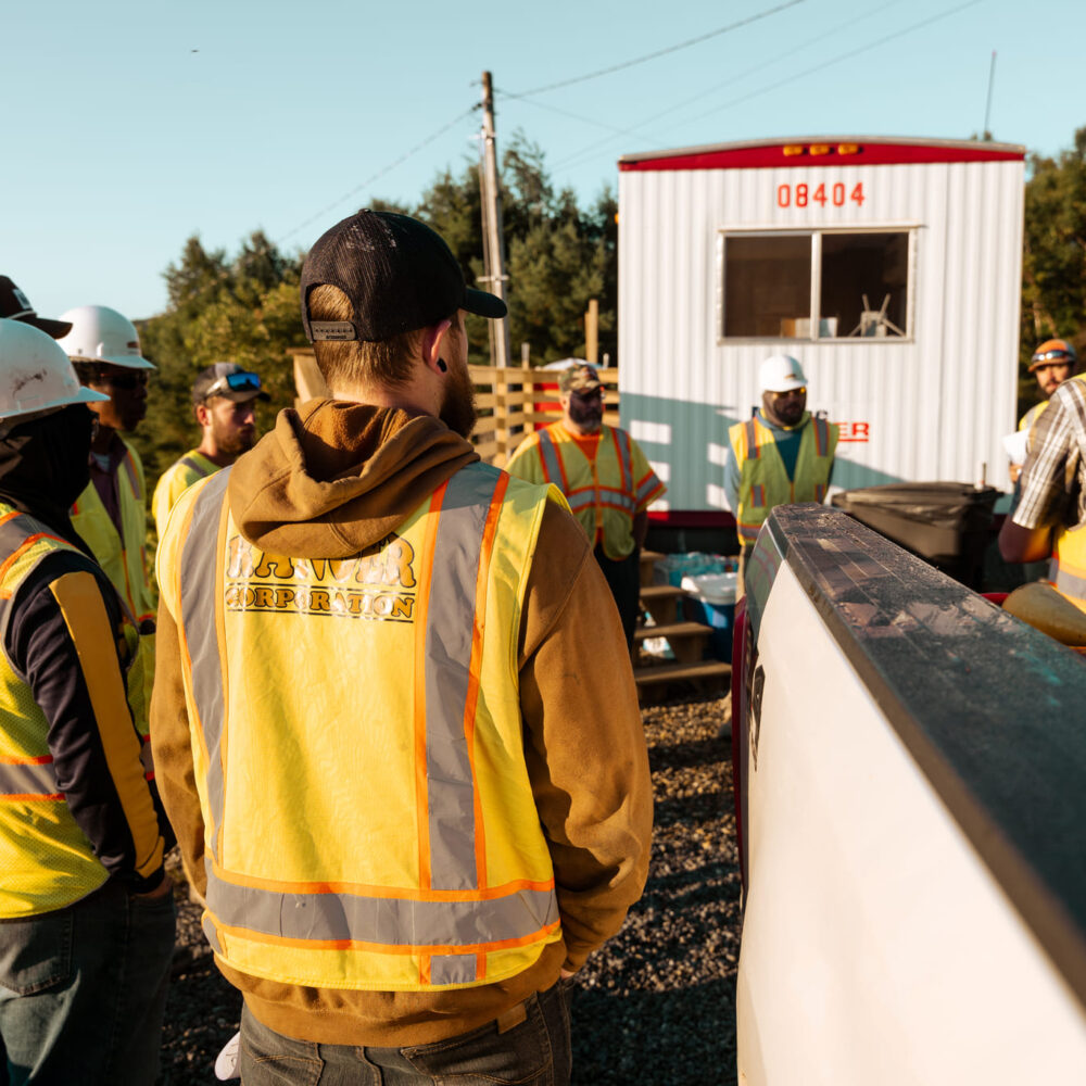 Construction workers in safety gear gather by a trailer outside.