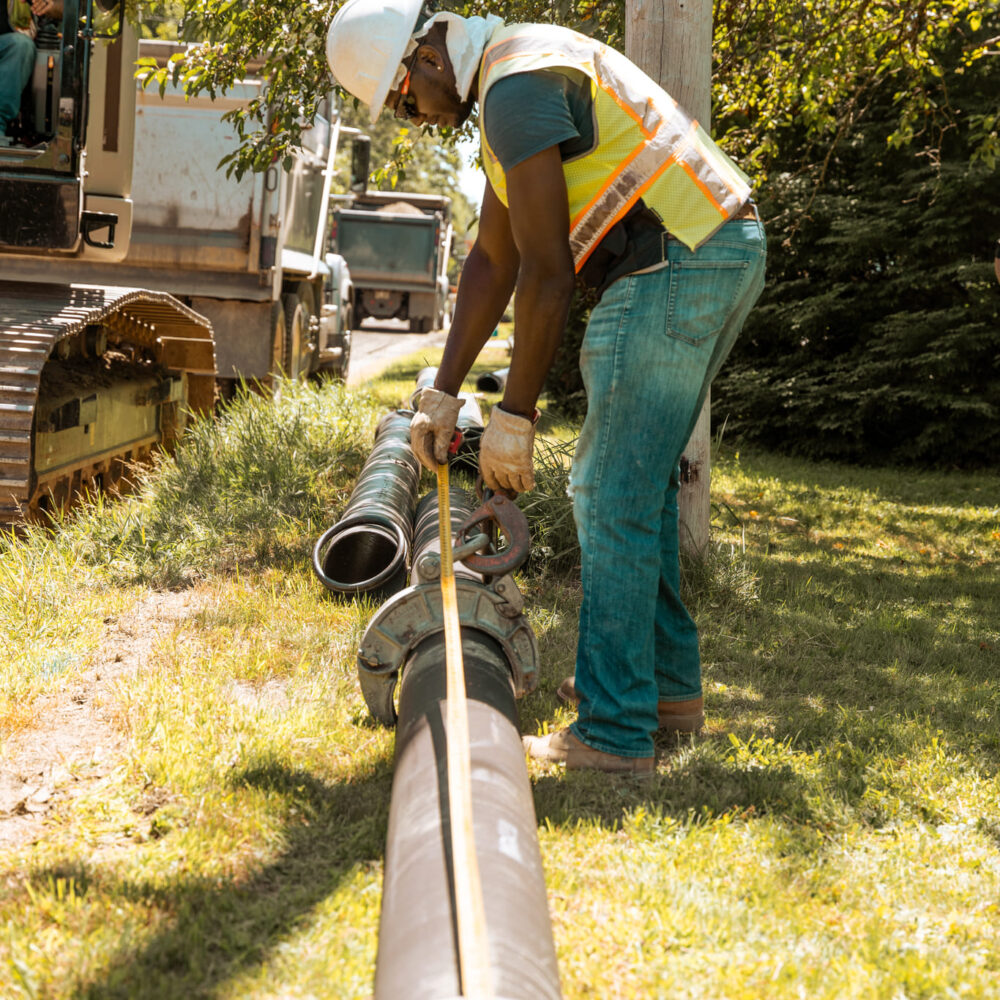 Worker in safety gear straps black pipe near machinery outdoors.