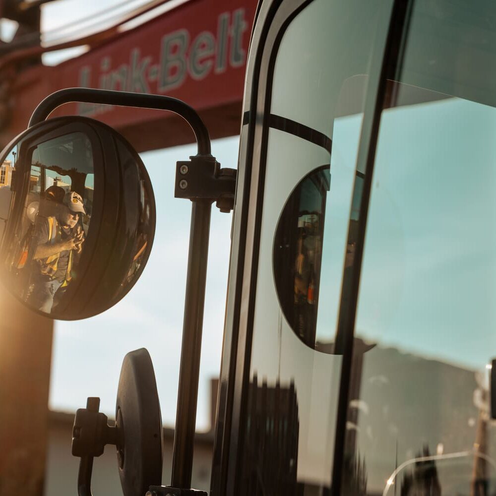 Side mirror reflects worker, vehicle parts, and sunlit crane.