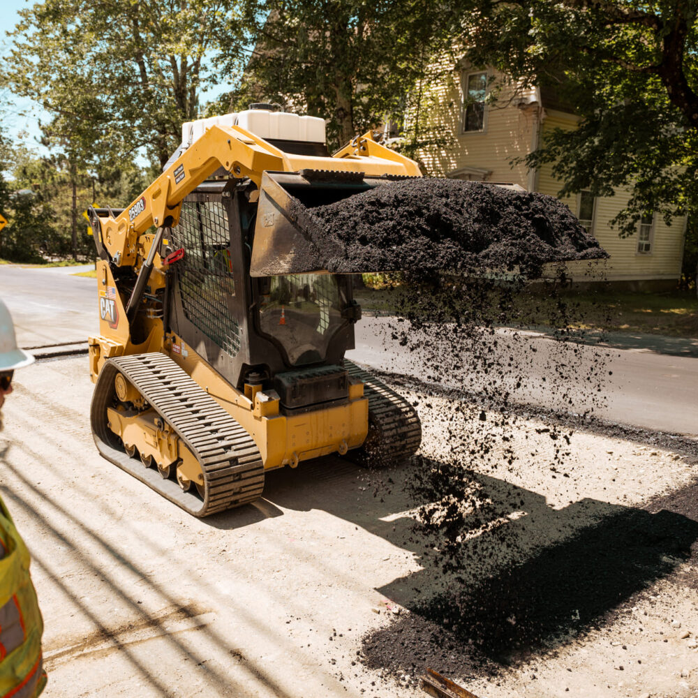 Worker watches yellow skid steer spread asphalt on sunny road.