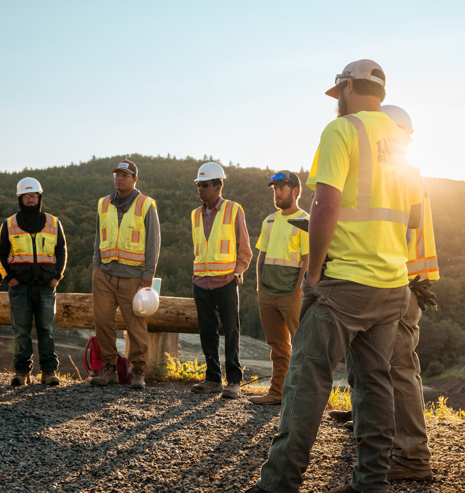 Six construction workers in vests and helmets stand on gravel.