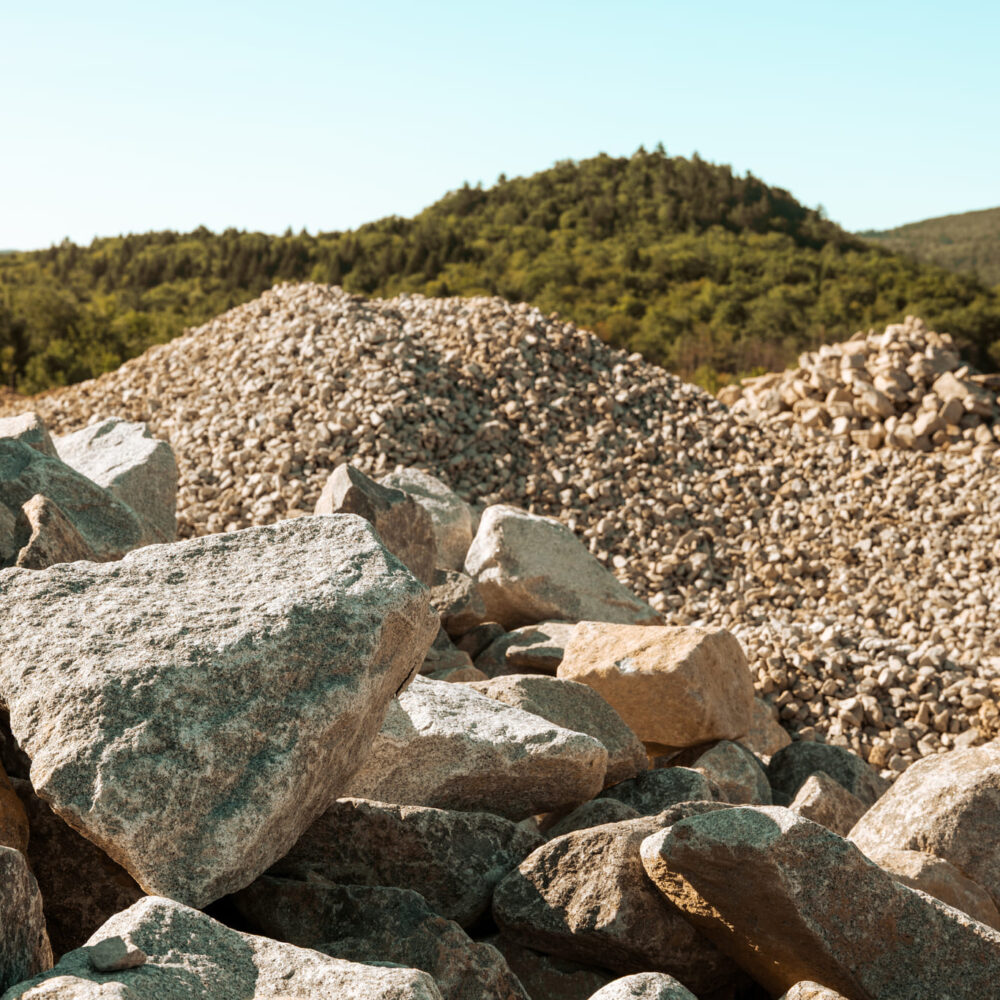 Rock and gravel piles outdoors with hills and trees behind.