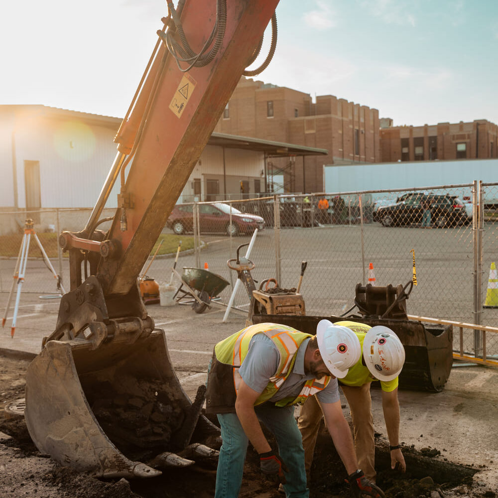Two construction workers inspect an asphalt hole by an excavator.