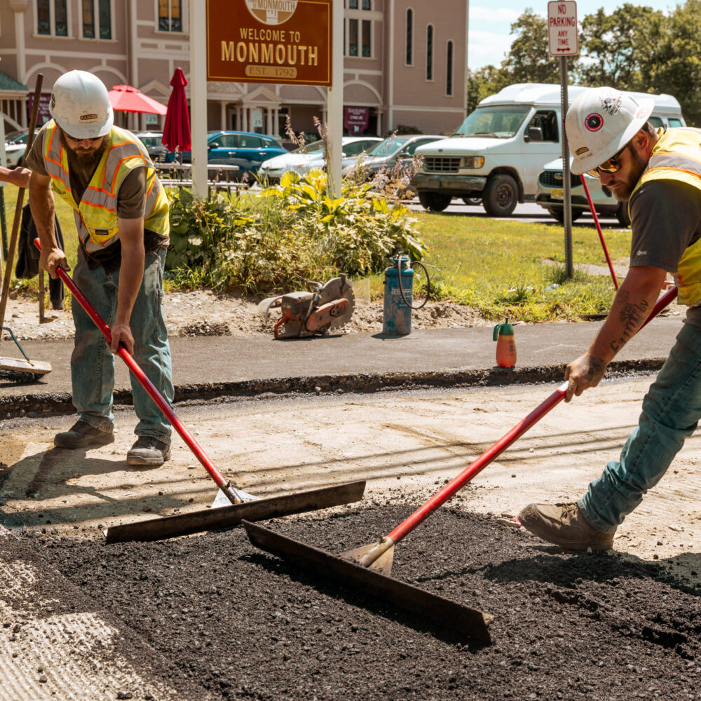 Workers spread asphalt by Welcome to Monmouth sign.