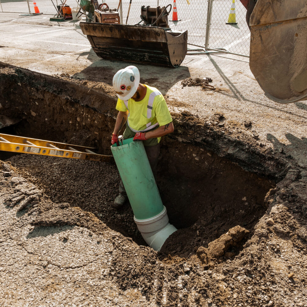 Worker in safety gear installs large green pipe in trench.