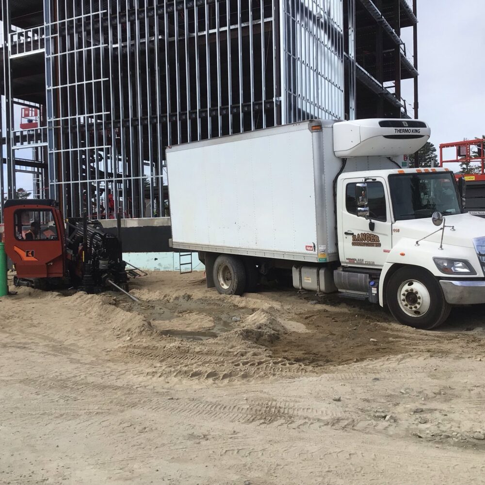 Box truck stuck in sand; excavator helps near steel-frame building.