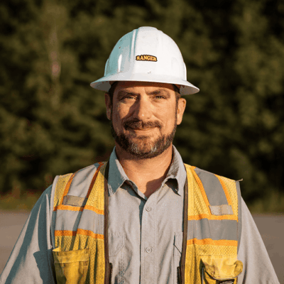 Construction worker in hard hat and safety vest outdoors by trees.