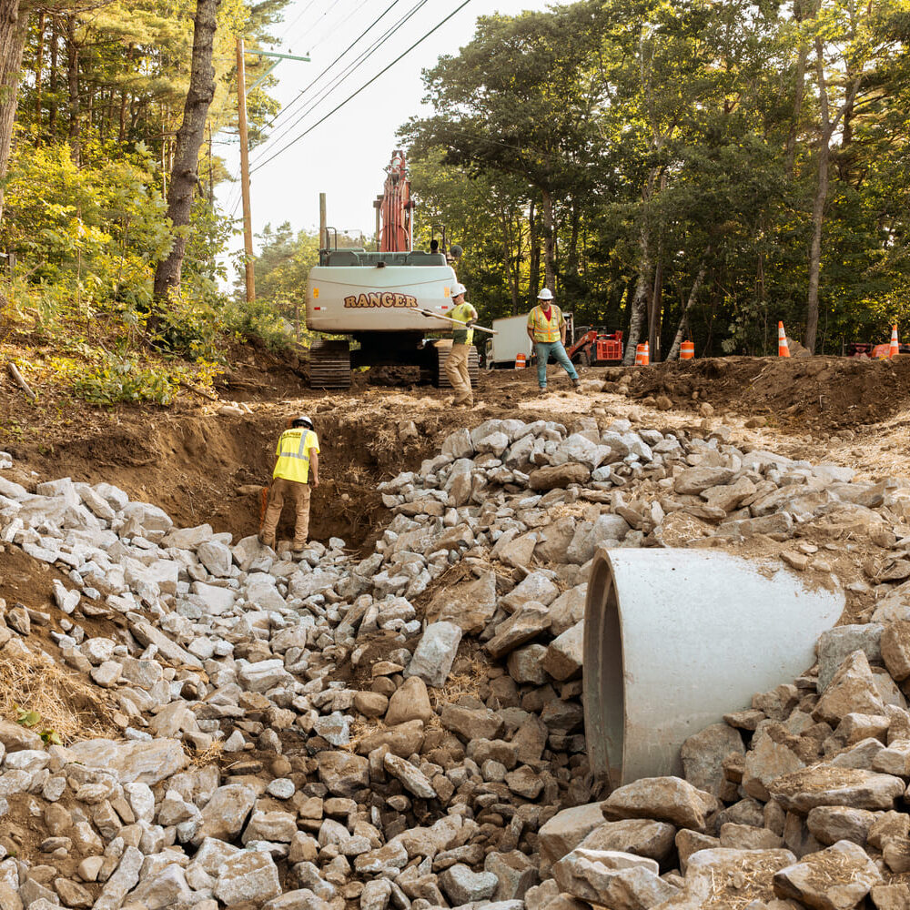 Workers by machinery, rocks, and a concrete pipe in the forest.