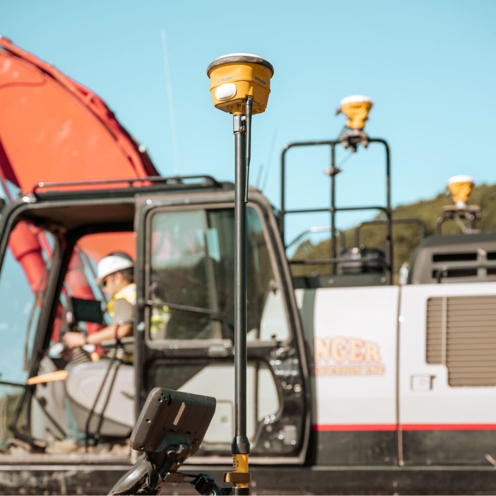 Yellow GNSS receiver on pole; worker with machinery behind.