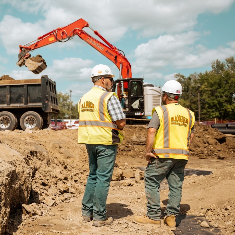 Workers in safety gear watch excavator load dump truck.