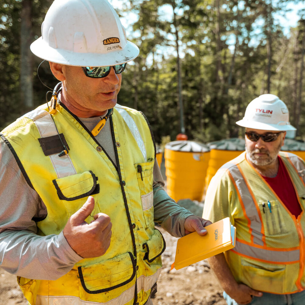 Two construction workers talk outside near large yellow containers.