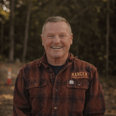 Smiling older man in plaid “Ranger Conservation” shirt outdoors.