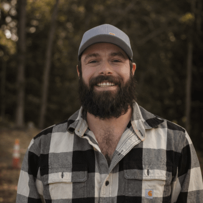 Bearded man in plaid shirt and gray cap stands outside.