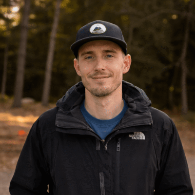 Man in black jacket and cap stands in wooded area.